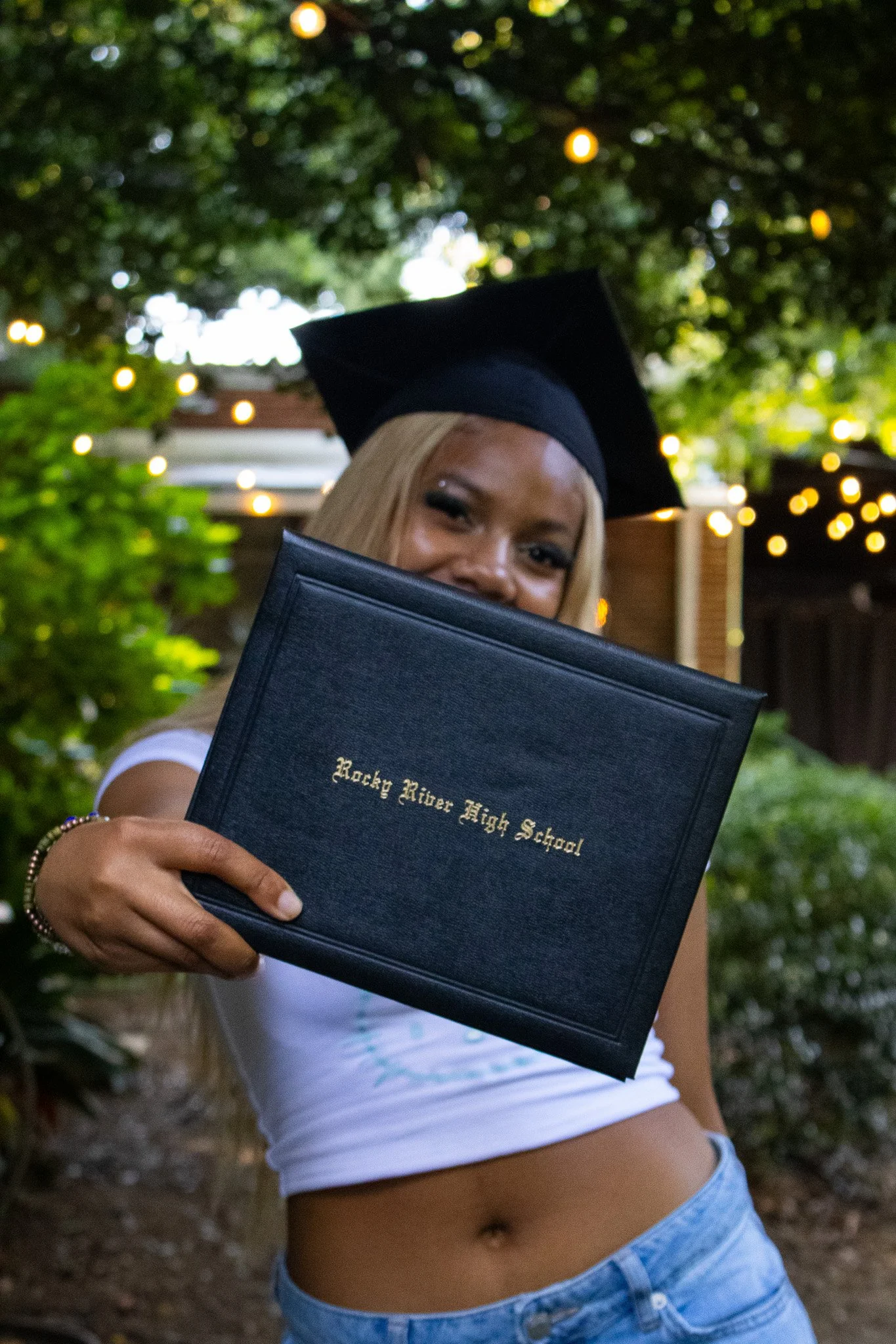 A young woman in a graduation cap holding a diploma cover from Rocky River High School, smiling outdoors with string lights and greenery in the background.