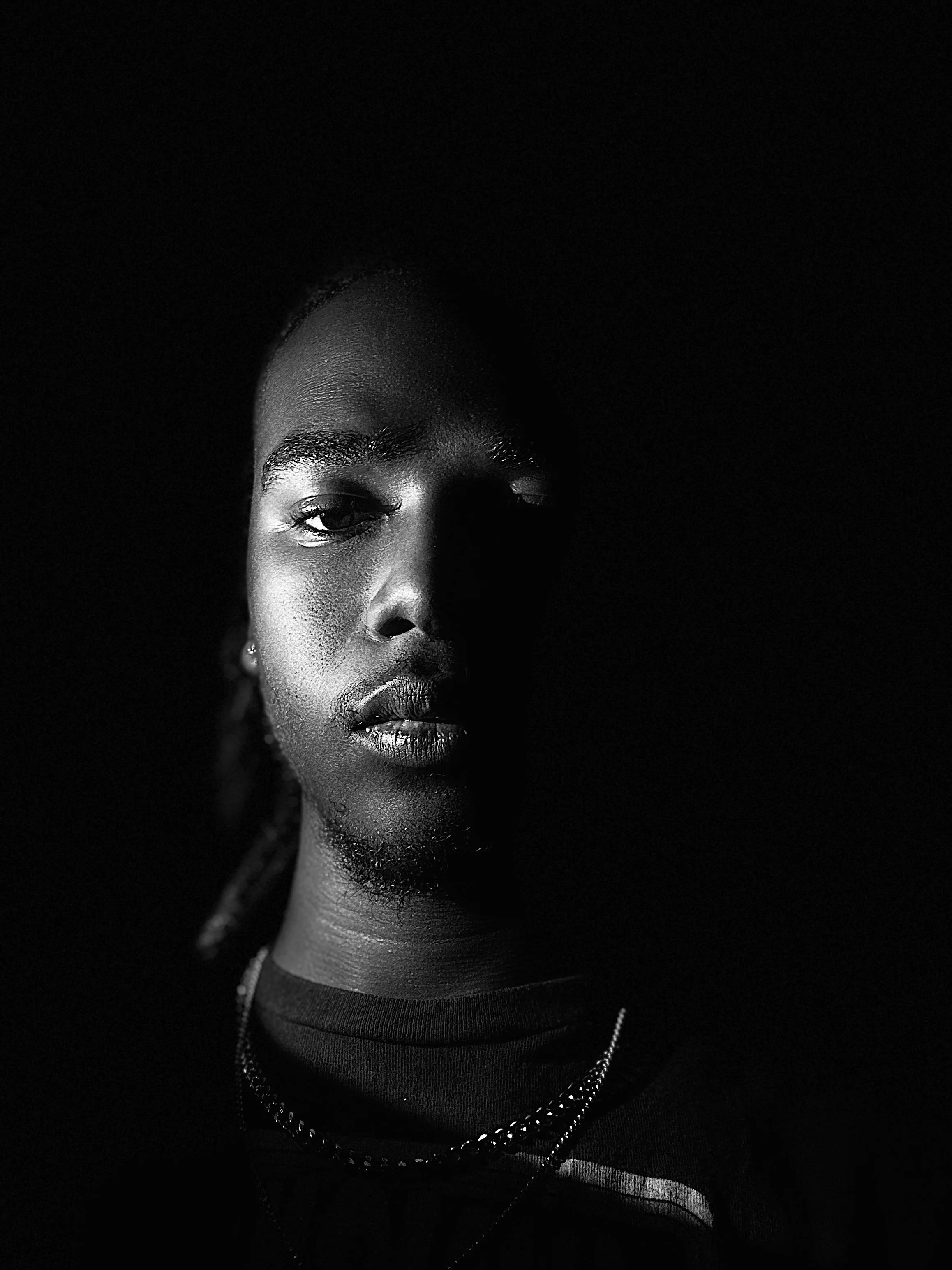 Black and white close-up portrait of a young woman with half of her face illuminated, featuring textured skin, earrings, necklace, and wearing a dark top.