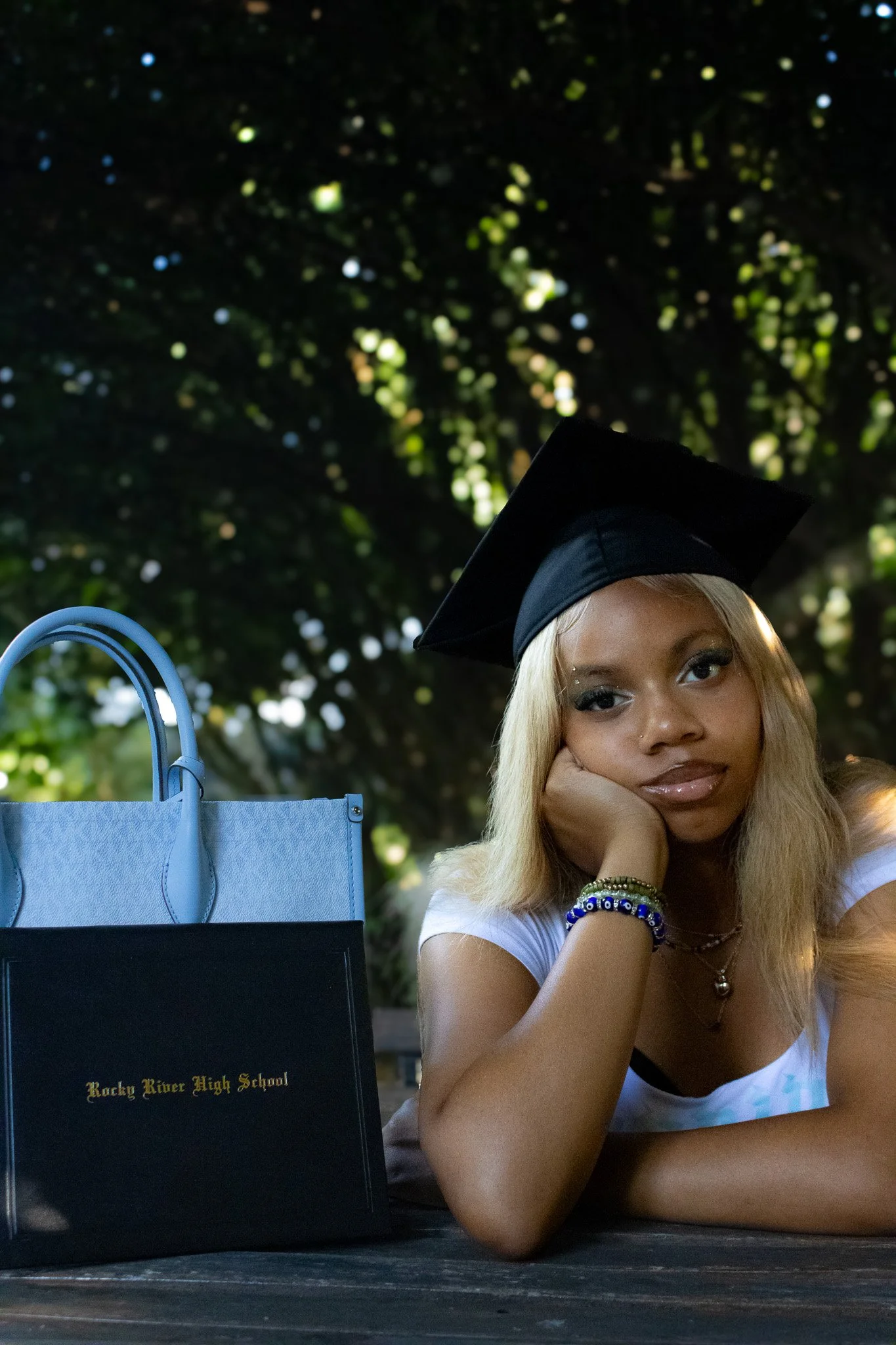 Young woman lying on a table outdoors, wearing a graduation cap, with Rocky River High School bag beside her, resting her face on her hand with trees in the background.