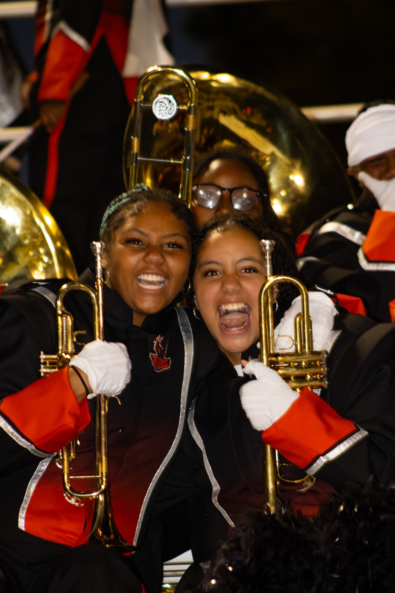 Two women holding brass instruments on a marching band float, wearing black and red band uniforms, excited and smiling during a celebration or parade.