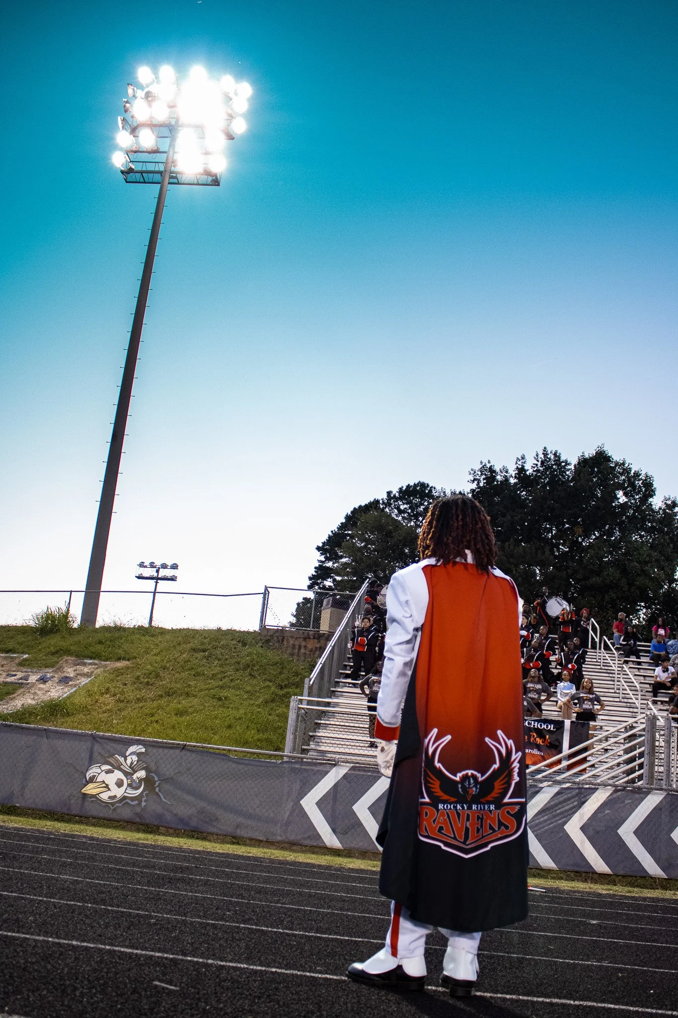 A person dressed in a band uniform with 'Rocky River Ravens' emblem on the back stands on a football field track, looking towards a stadium filled with band members and spectators, under a bright stadium light on a clear evening.