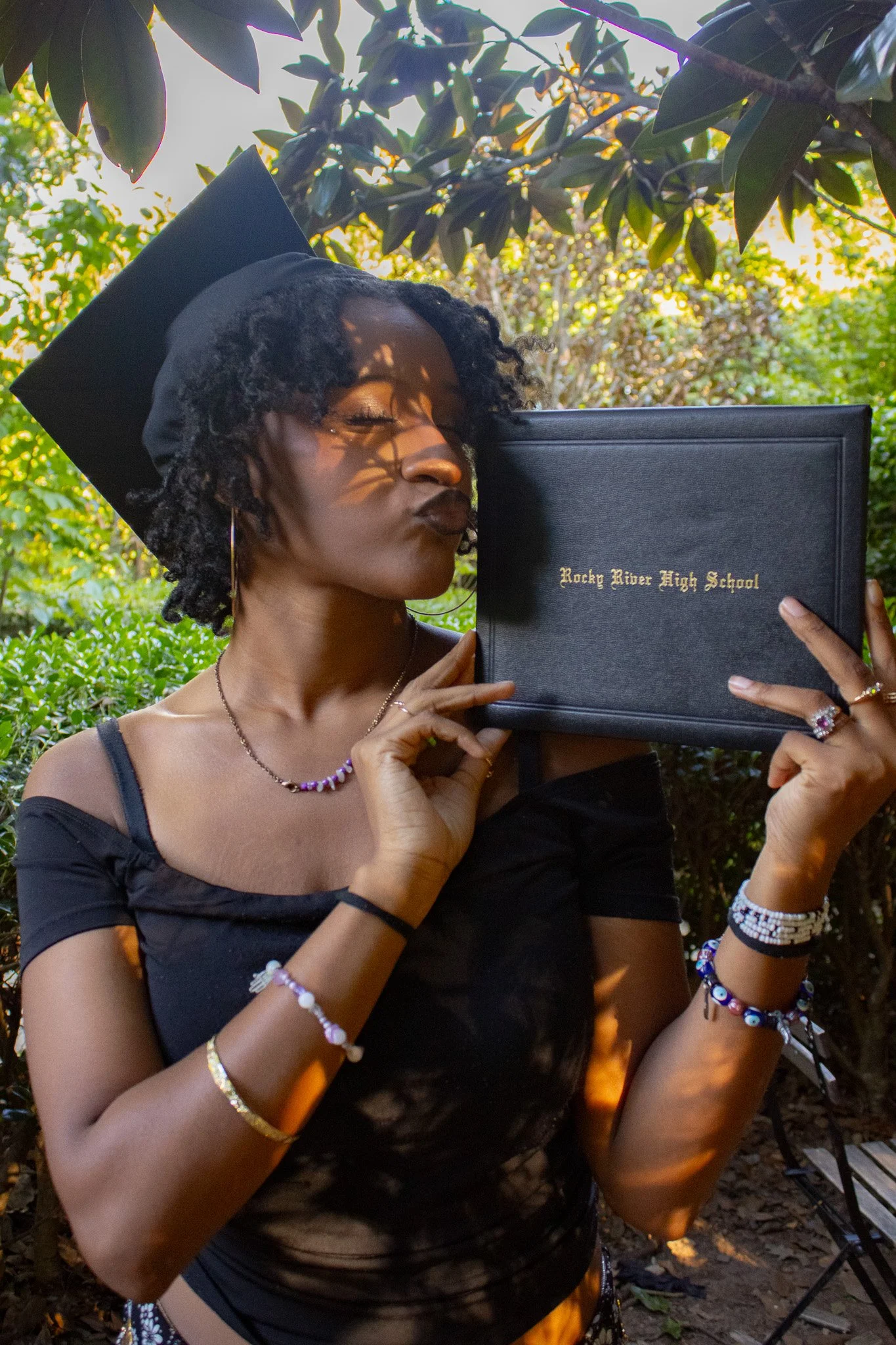 A woman in a graduation cap and black dress holds a diploma from Rocky River High School close to her face outdoors amidst greenery, making a kissing gesture.