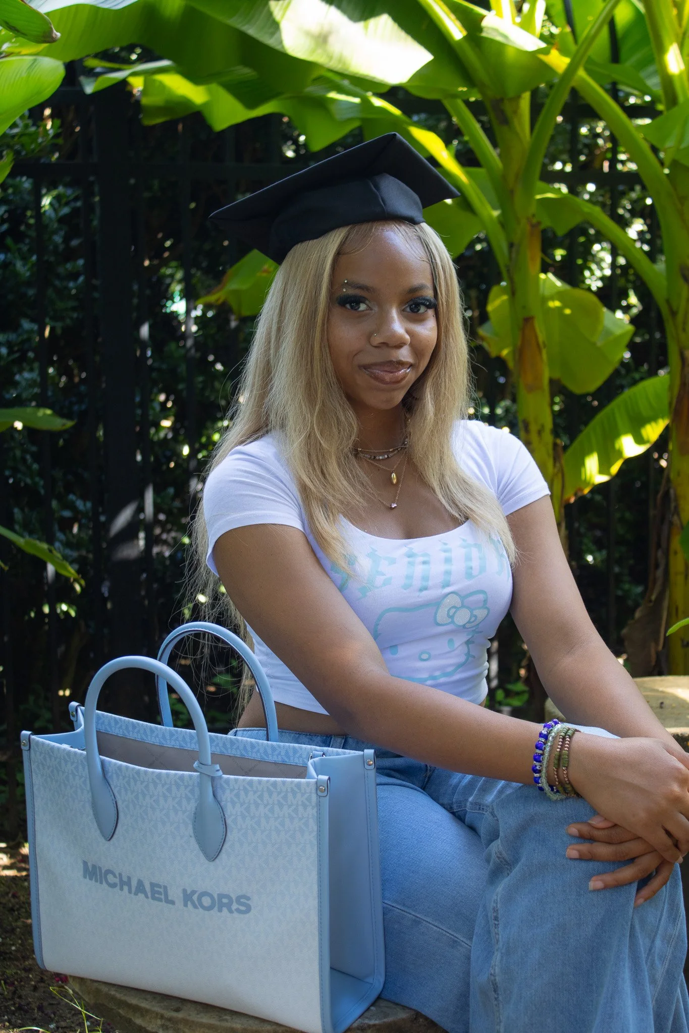 A young woman wearing a graduation cap sits outdoors surrounded by green plants, with a light blue Michael Kors tote bag nearby.