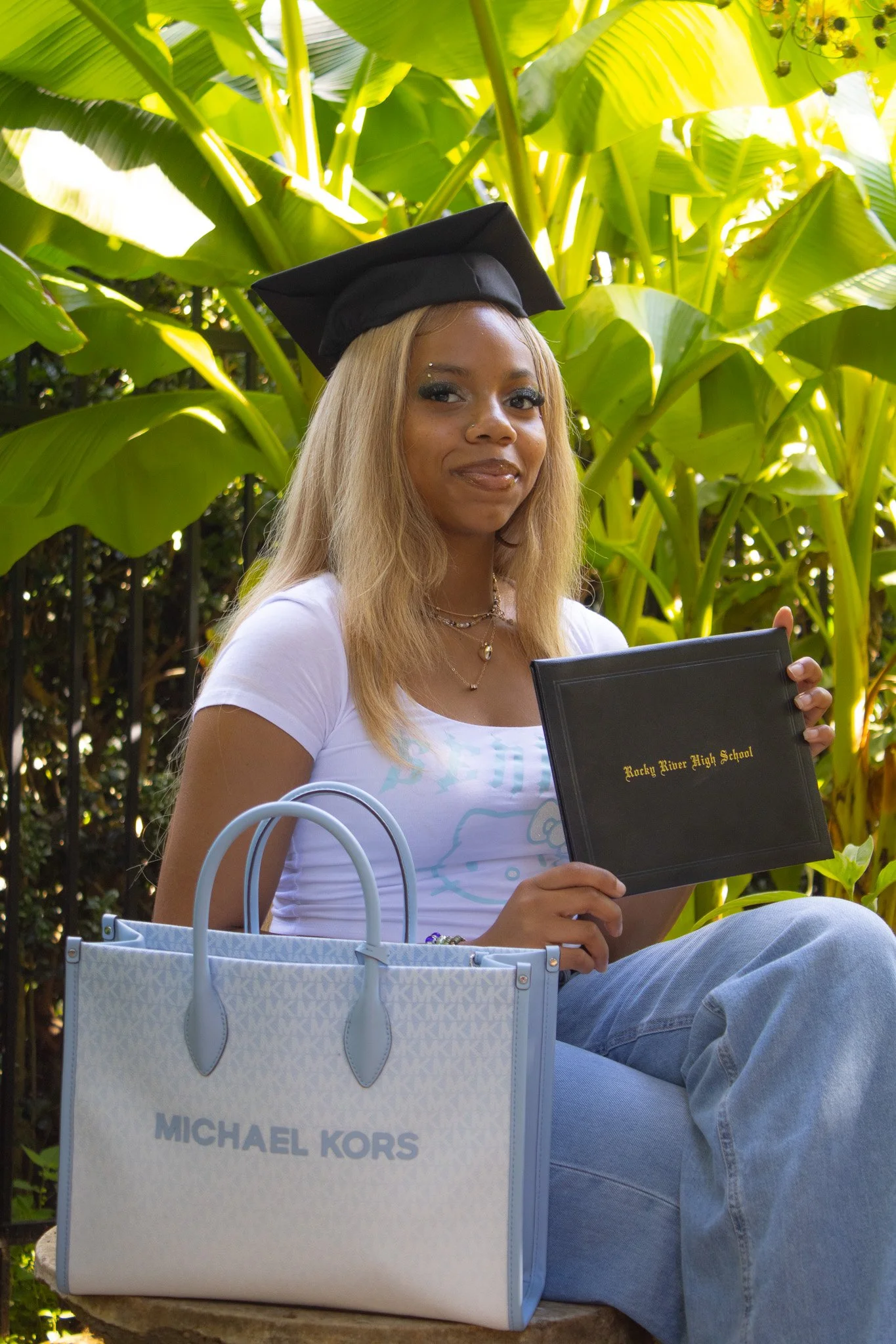 A young woman with blonde hair and a graduation cap, sitting outdoors among lush green plants, holding a black diploma folder from Rocky River High School, with a white Michael Kors tote bag beside her.