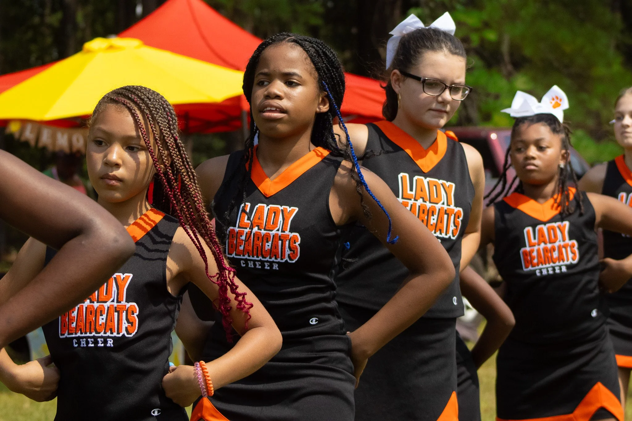 Cheerleaders from Lady Bearcats team standing outdoors with their hands on hips, wearing black and orange uniforms with white bows in hair, in formation during a cheer event.