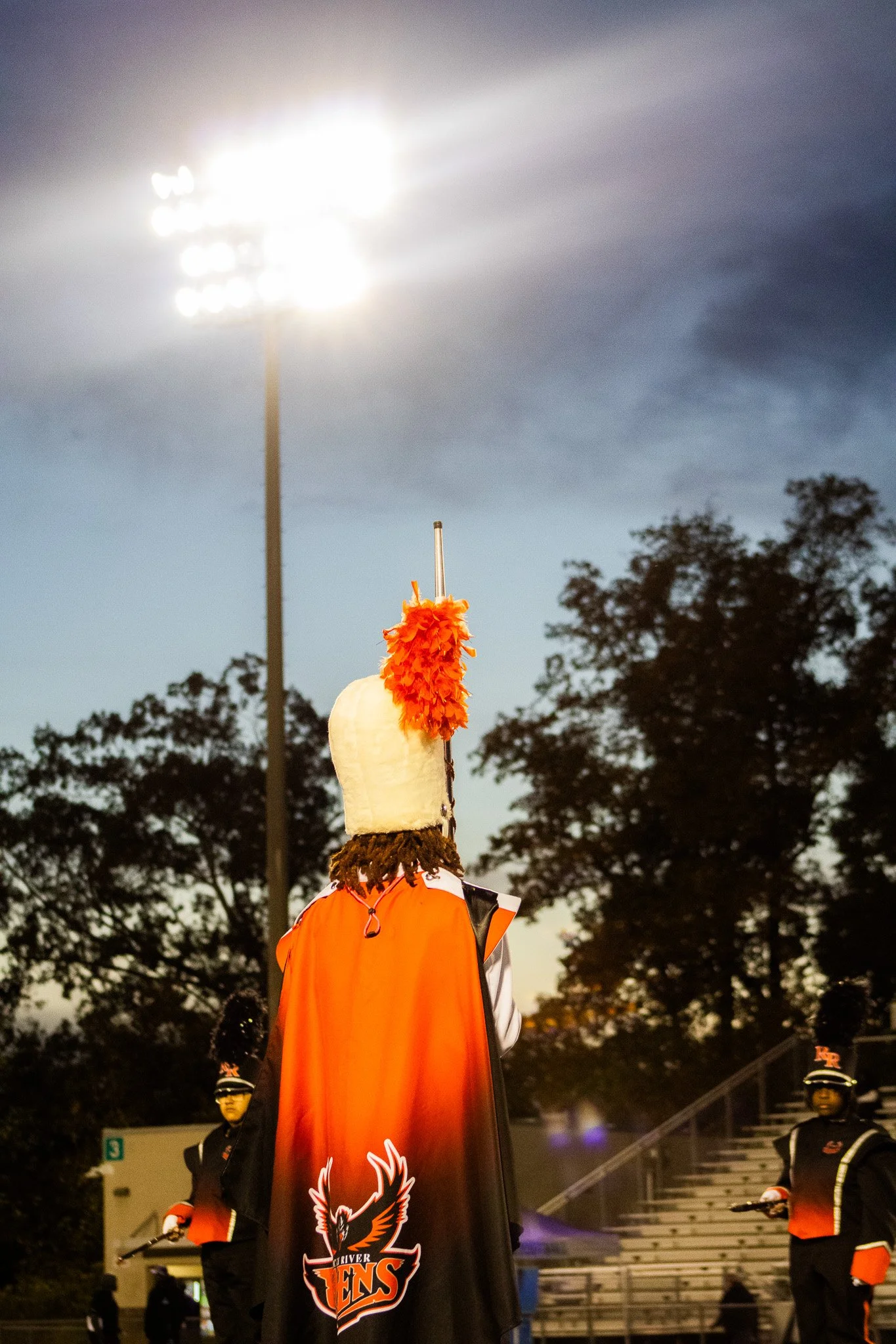 A marching band performer in a black and orange uniform with a cape featuring a Phoenix logo, wearing a tall, white hat decorated with orange feathers, standing outdoors at dusk with trees and stadium lights in the background.