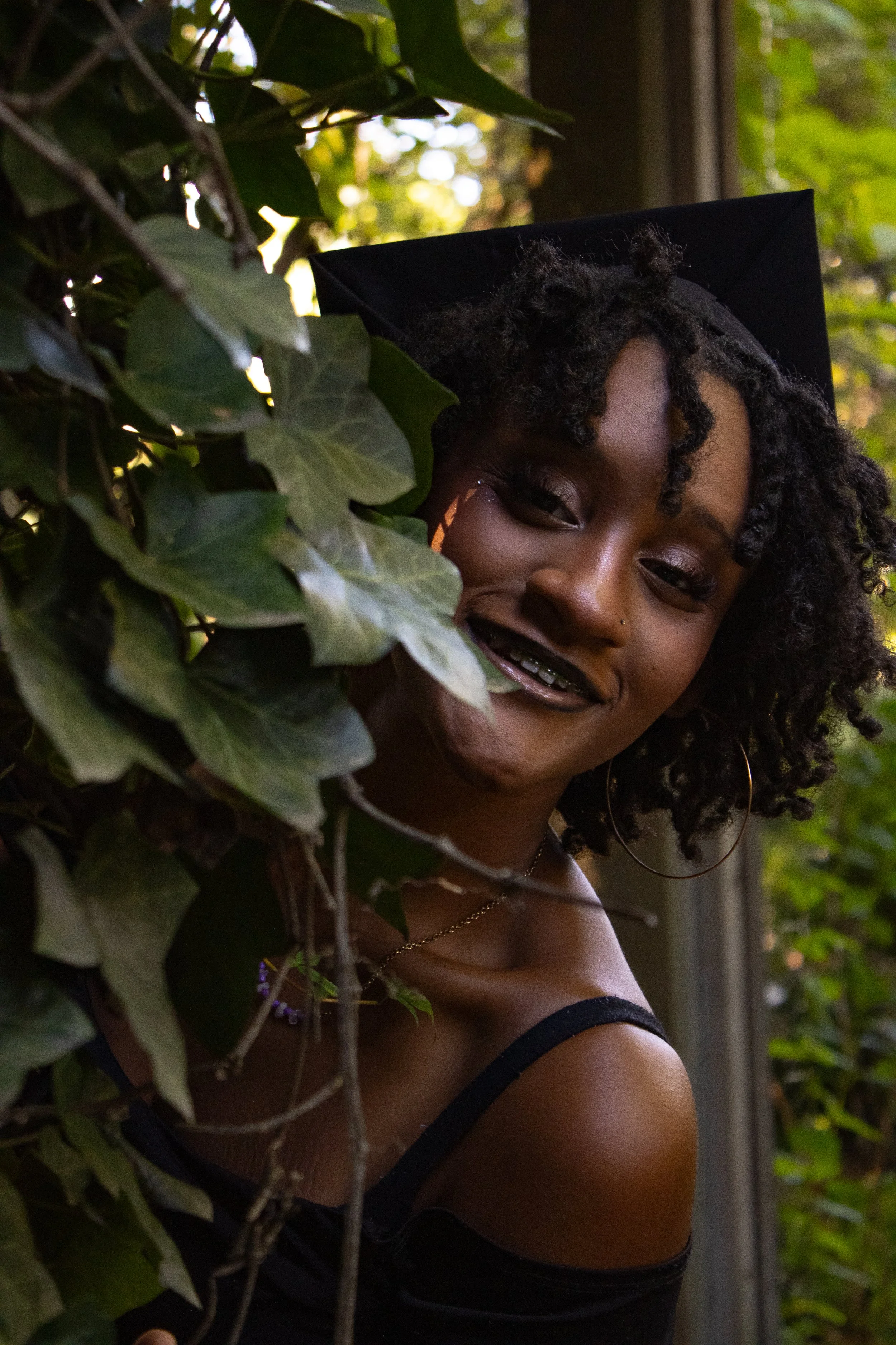 A woman with curly hair and hoop earrings sporting a black off-shoulder top, smiling while peeking through greenery with sunlight filtering through leaves.