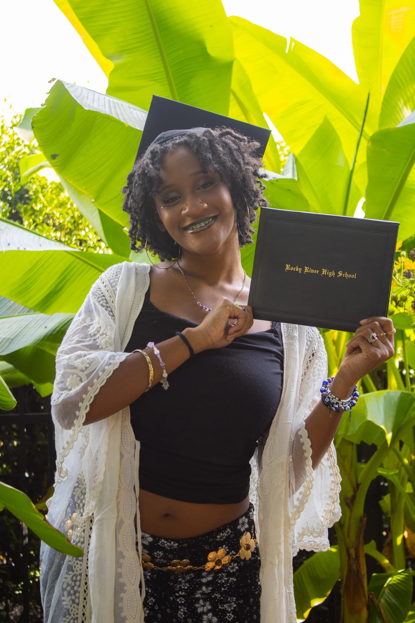 A young woman in a graduation cap and gown smiling and holding a diploma in front of green tropical leaves.