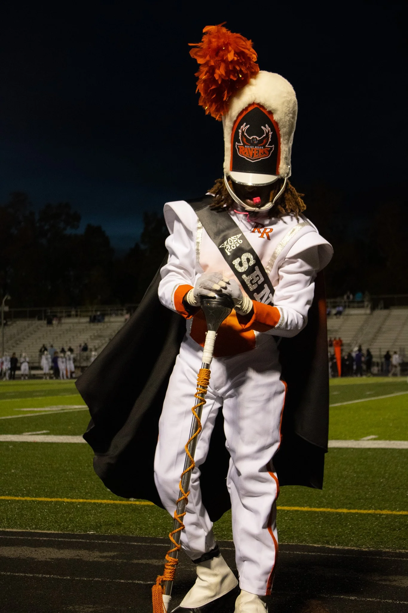 A person dressed in a football referee costume with a marching band hat that has a Raven's logo, a cape, and a sash that reads 'HOME TEAM'. The individual is holding an orange and white baton on a football field during nighttime.