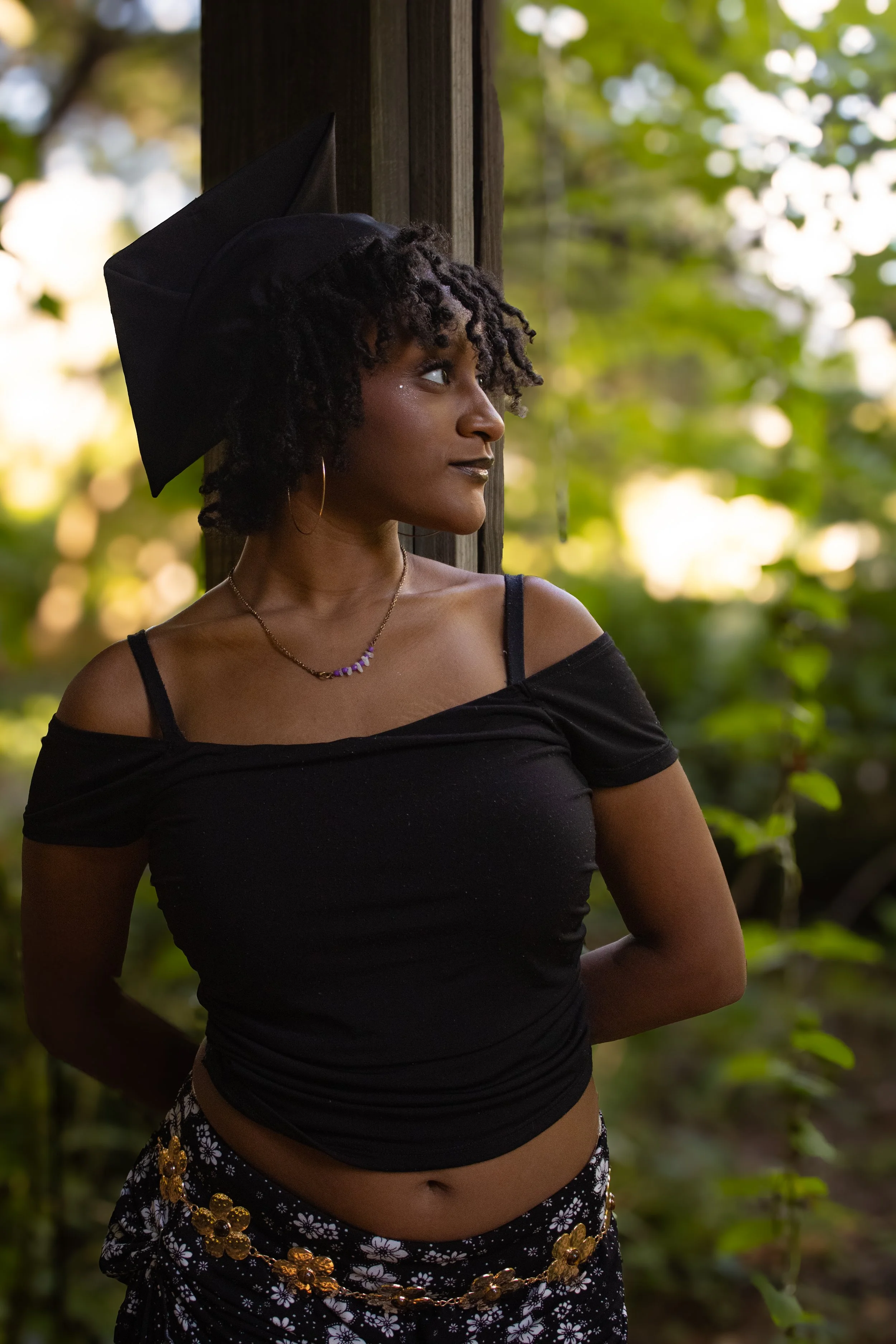A young woman in a graduation cap looking to the side outdoors with trees in the background.