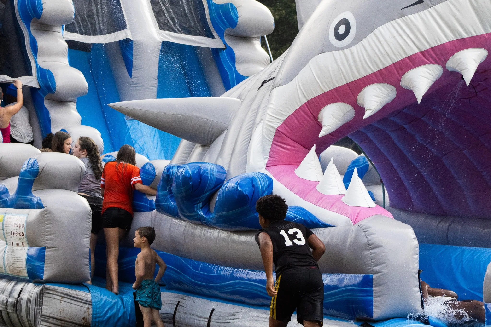 Kids and adults on a large inflatable water slide designed to look like a shark with its mouth open, white teeth, and pink gums. Water is splashing as they slide down.