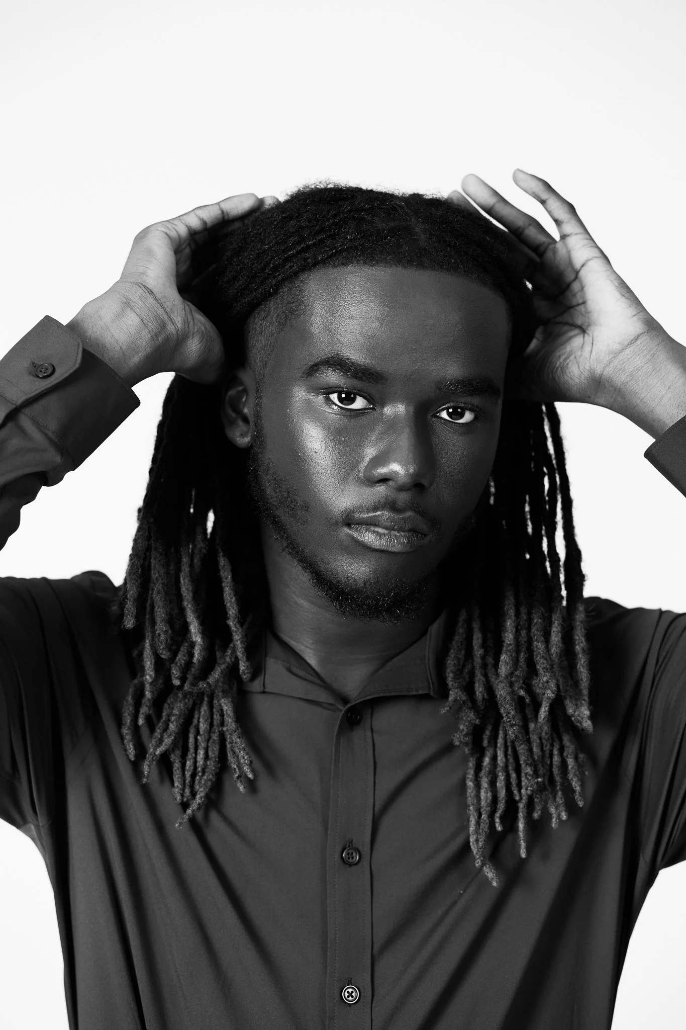 A black and white portrait of a young man with long dreadlocks, wearing a button-up shirt, looking directly at the camera with his hands behind his head.