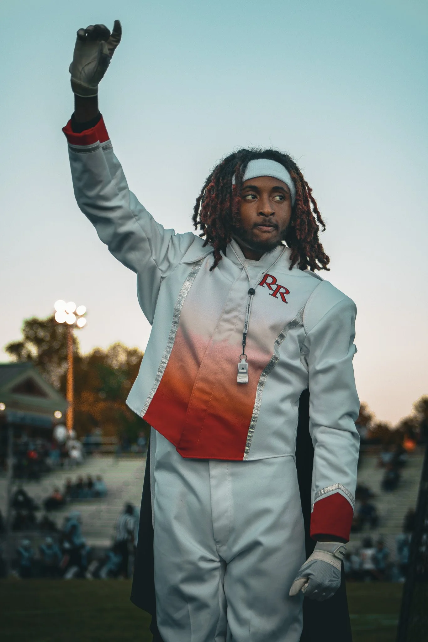 A person dressed in a racing suit with the initials 'RR' raises their arm on a race track at dusk, with an audience in the background.