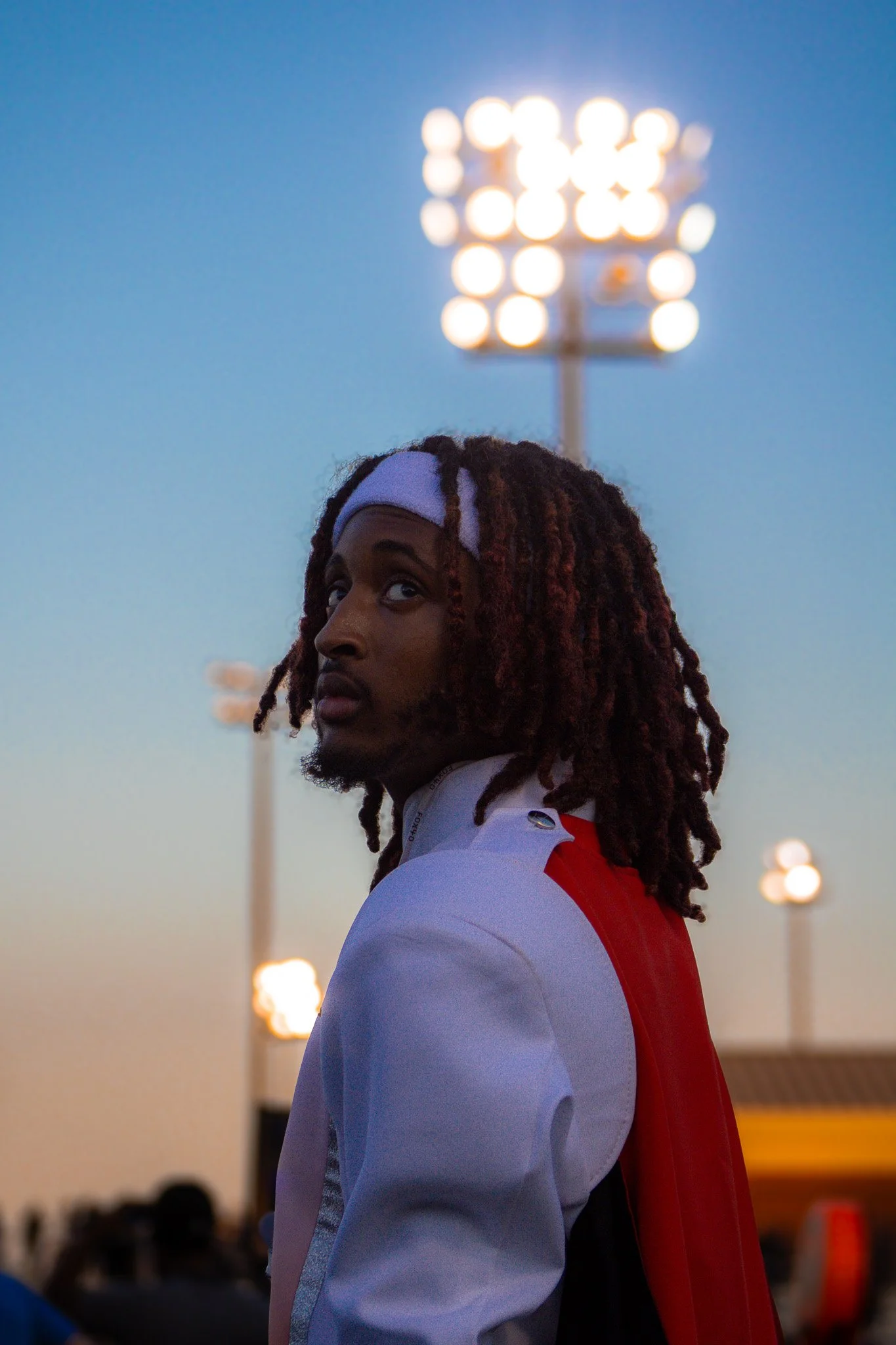 A man with long, curly dreadlocks wearing a white headband and a jacket with white, red, and black colors standing outdoors during dusk with stadium lights in the background.