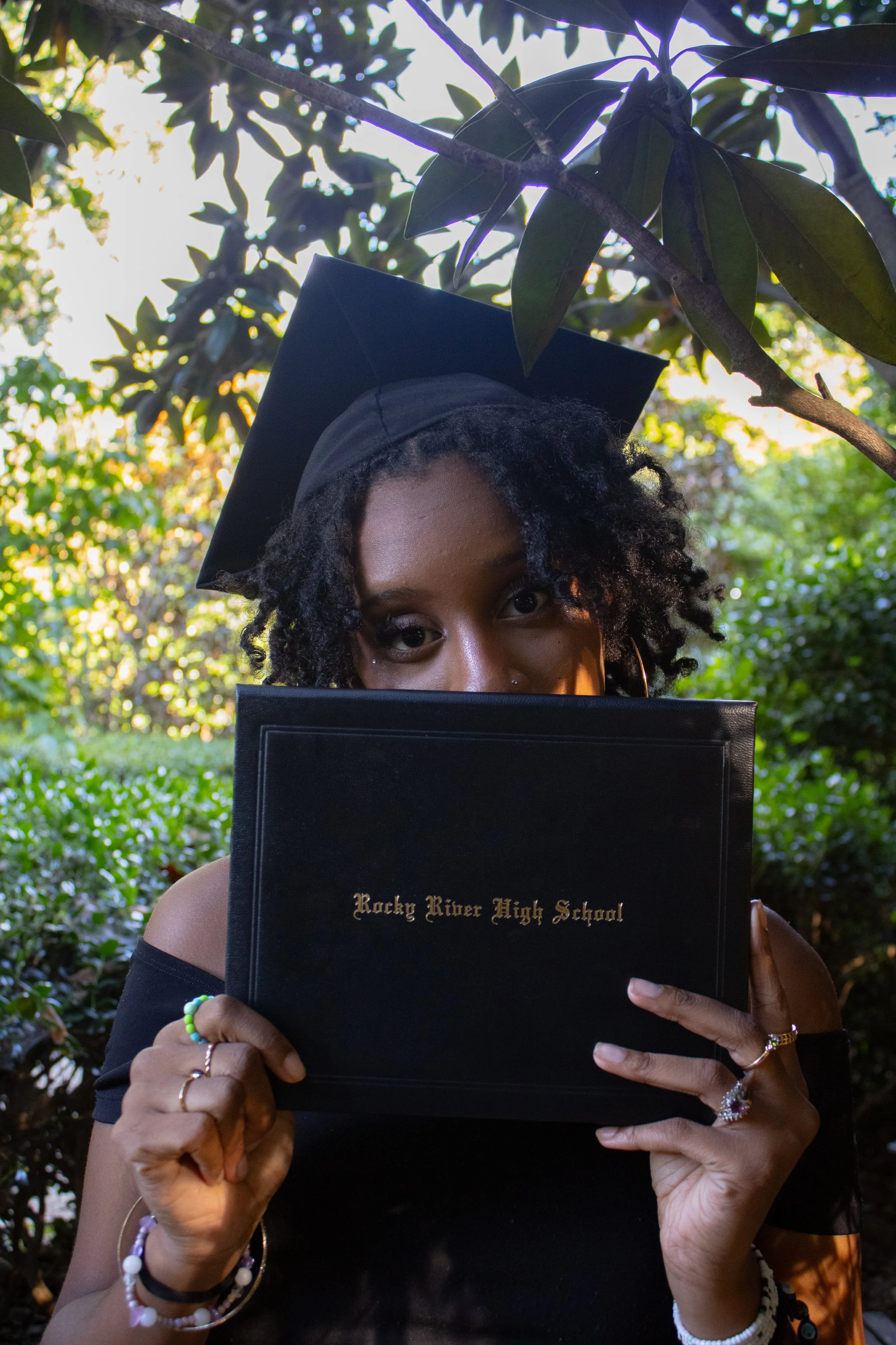 A young woman wearing a graduation cap and holding a diploma boat with the text 'Rocky River High School' in front of her face, outdoors among trees and greenery.