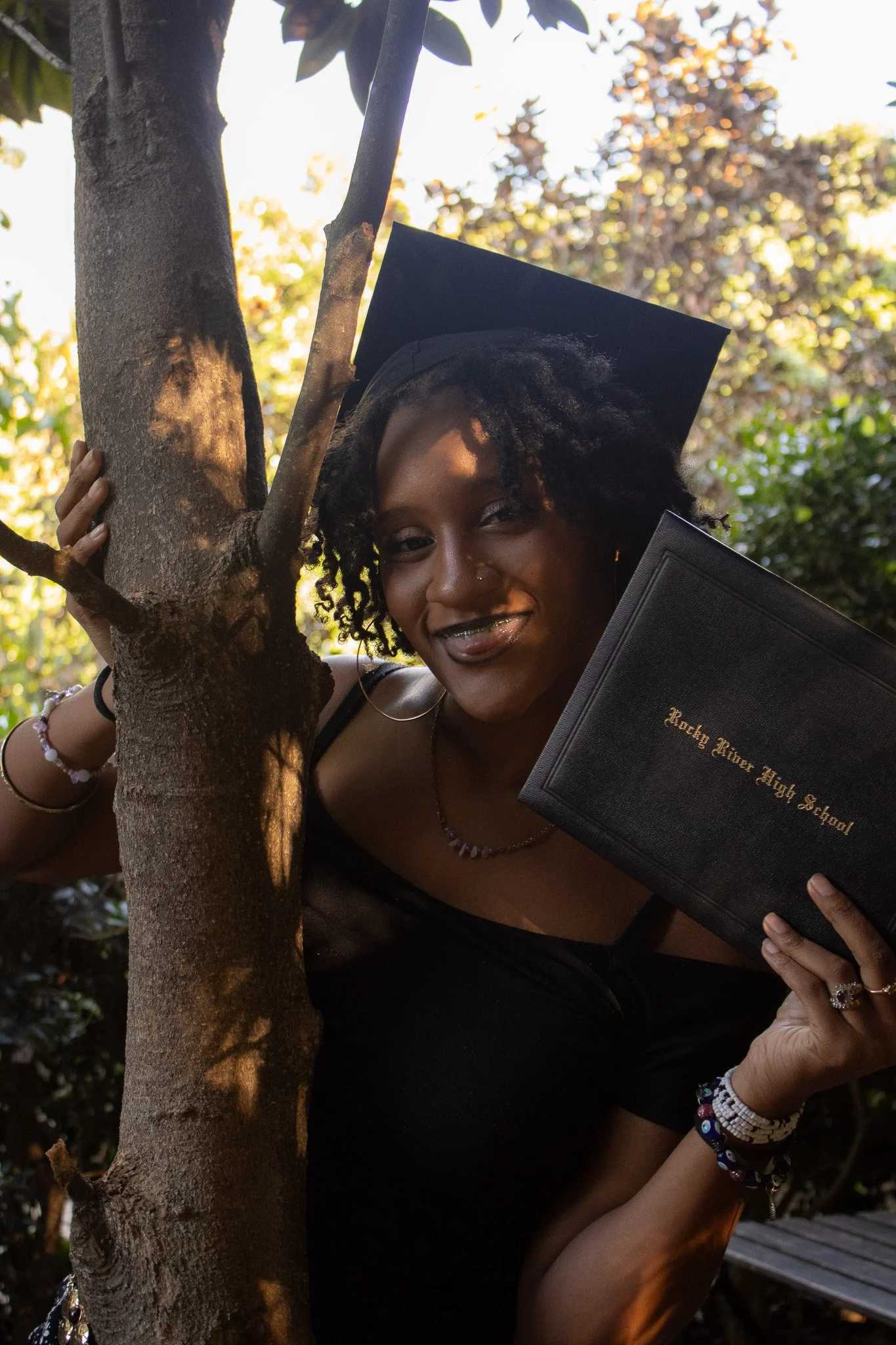 A young woman in a graduation cap and gown holding a diploma while peeking out from behind a tree, outdoors with sunlight filtering through the leaves.