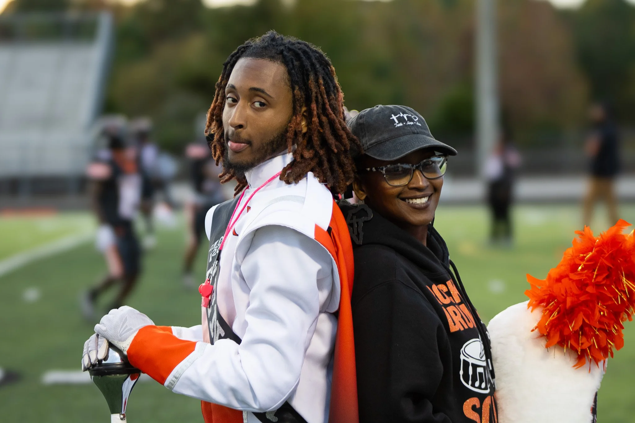A young man and woman standing back-to-back on a football field, smiling. The man is dressed in a white football uniform with an orange stripe and gloves, holding a helmet. The woman is wearing glasses, a black hoodie, and a black cap, holding an ora