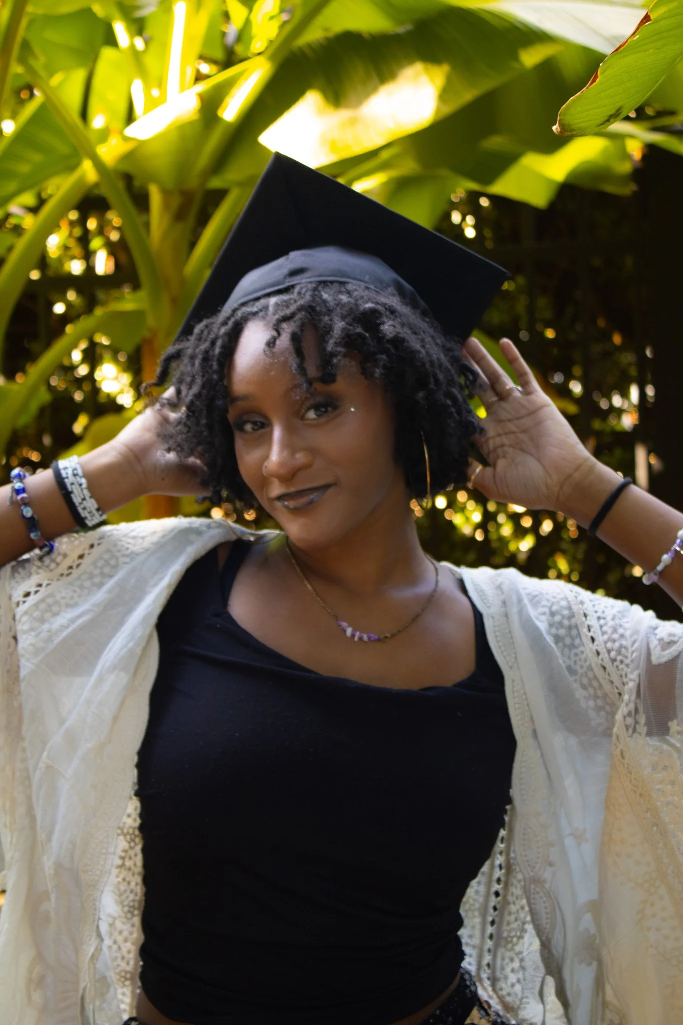 A young woman in a graduation cap and gown poses outdoors among large green leaves.