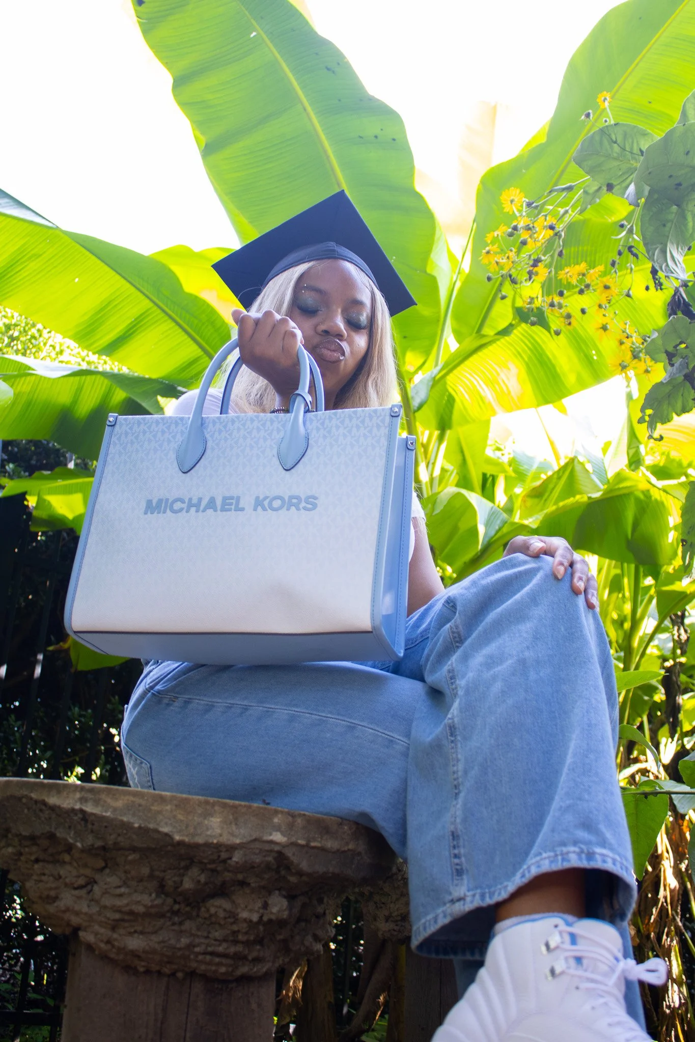 Young woman wearing a graduation cap, sitting outdoors on a wooden bench, holding a large Michael Kors handbag, with lush green tropical plants behind her.