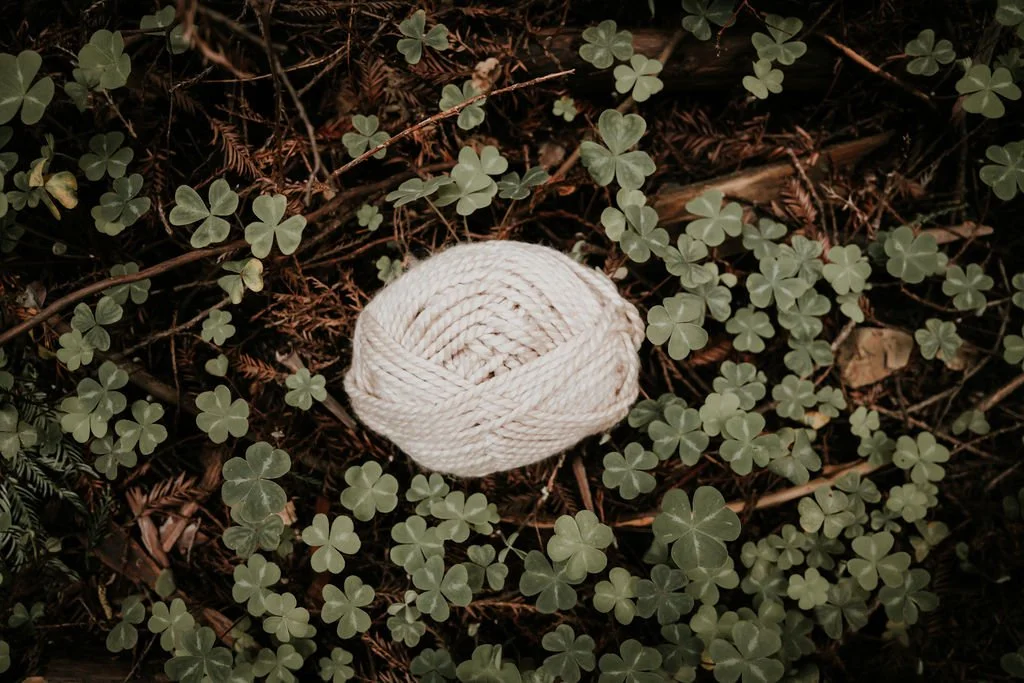 A ball of white yarn placed on a bed of green clover and brown plant debris.