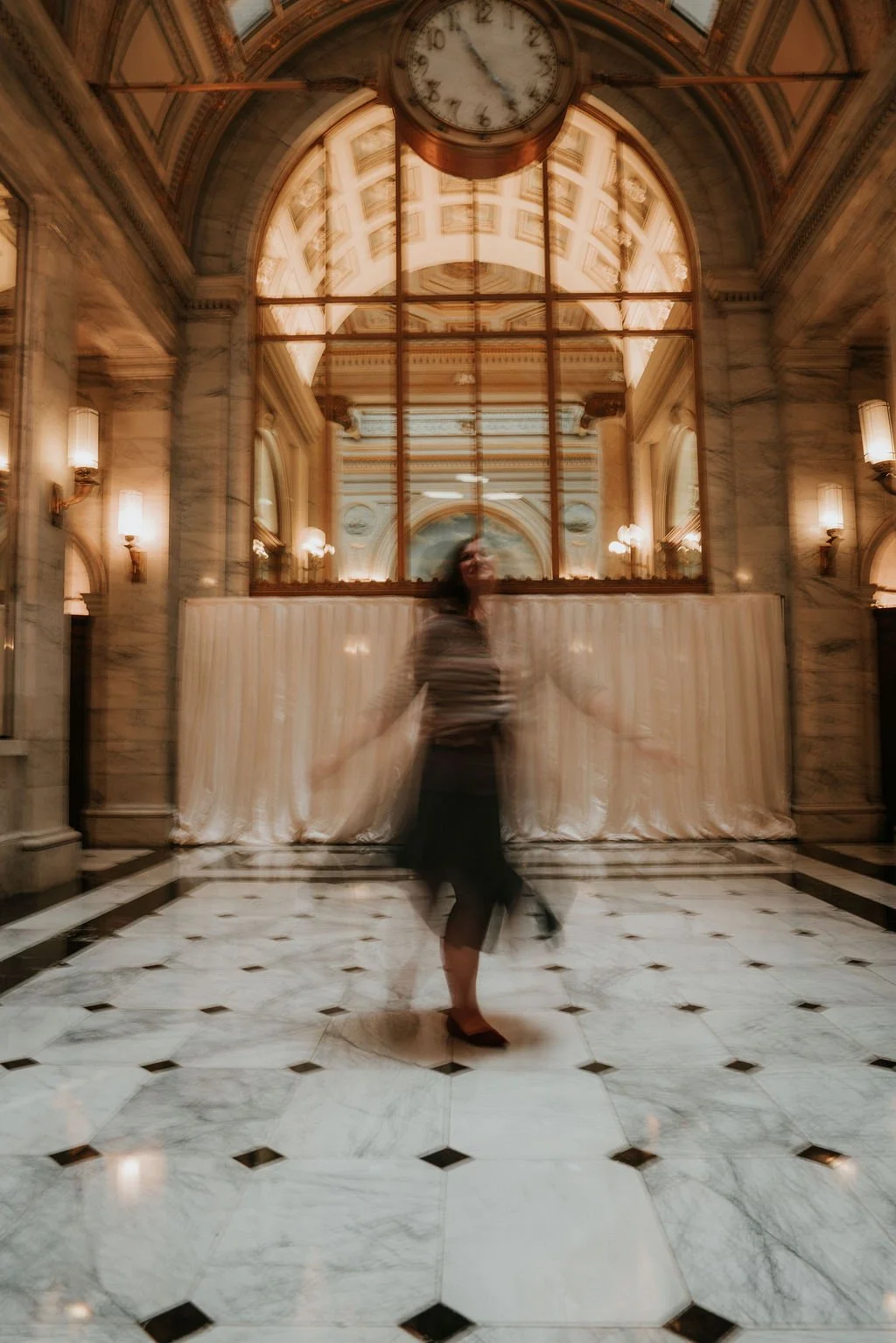 Blurry photo of a woman dancing or spinning inside an elegant, historic building with marble floors, ornate walls, large clock, and high, arched windows reflecting the ceiling.
