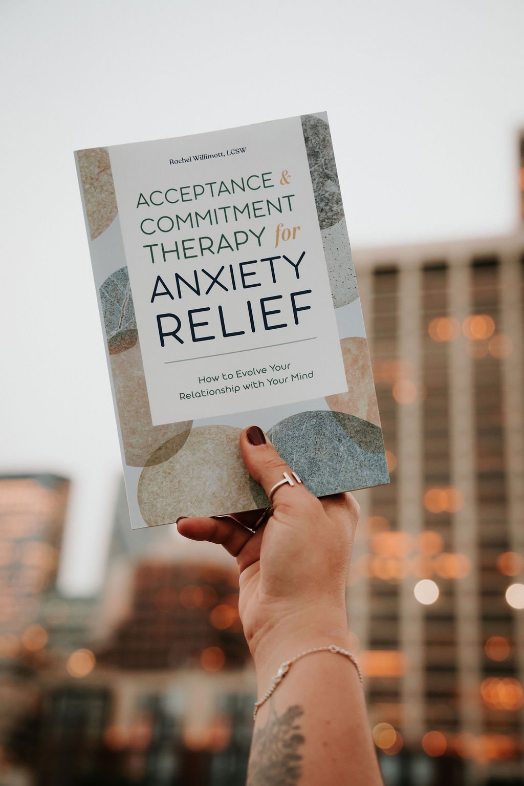 Person holding book titled 'Acceptance & Commitment Therapy for Anxiety Relief' with city buildings and lights in the background.