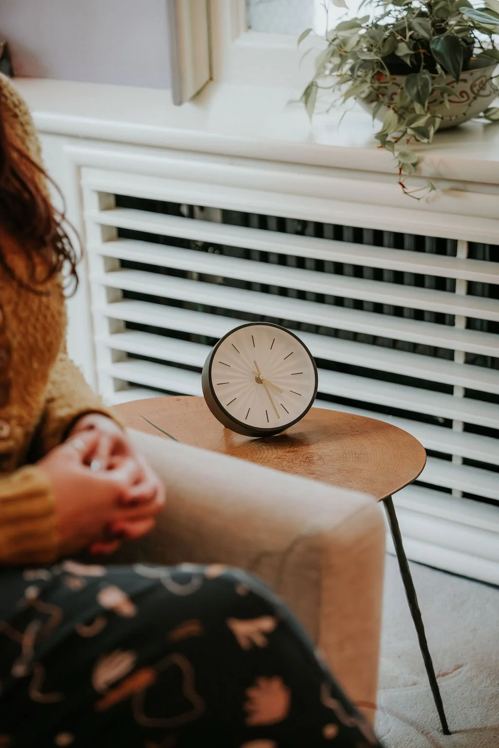 A round modern clock with black frame and light face displaying time, placed on a wooden table next to a white radiator and a potted plant.