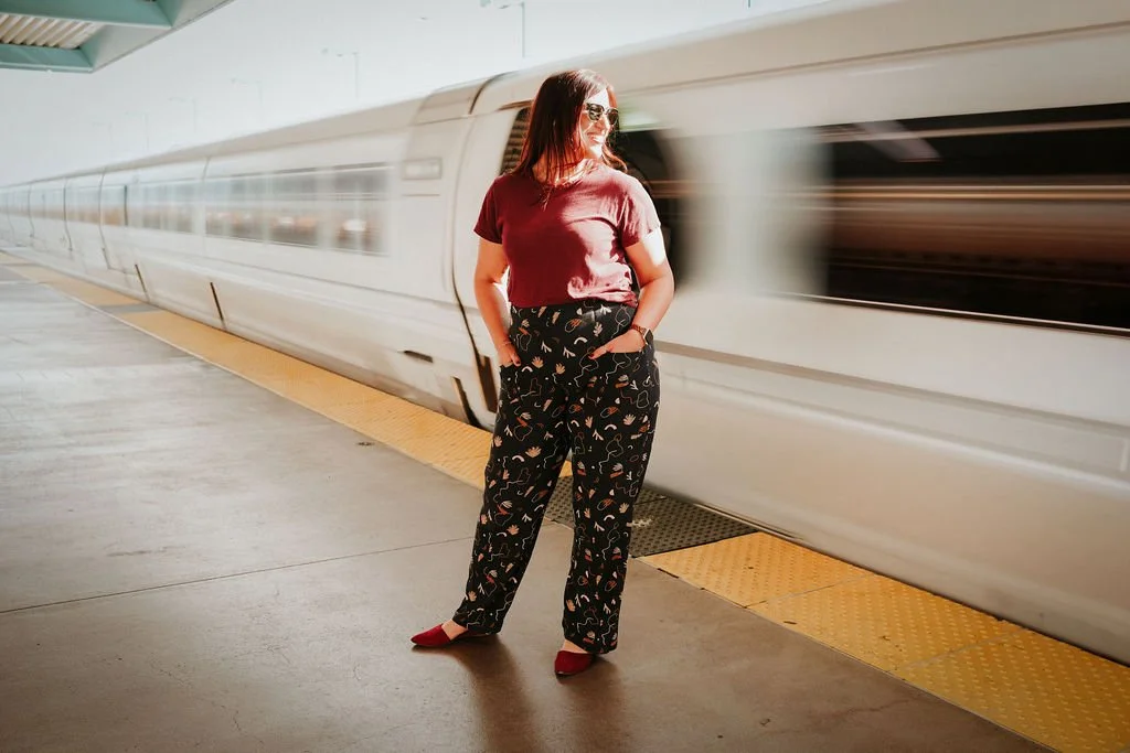 A woman wearing sunglasses, a maroon T-shirt, and patterned black pants, standing on a train platform with a high-speed train passing by.