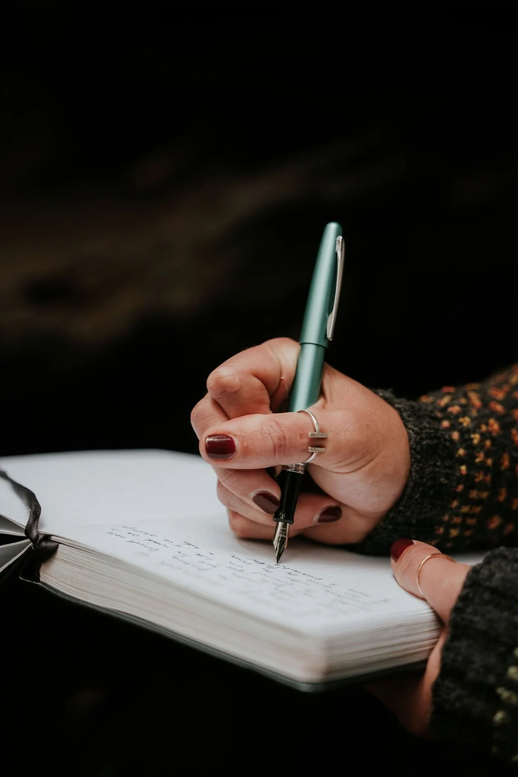 Close-up of a person's hand holding a mint green pen writing in a black notebook with a ribbon page marker, dark background.