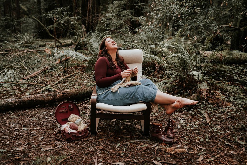 A woman with long hair and a maroon top is sitting on a white chair in a forest, laughing while knitting. Her shoes are off and on the ground beside her. There is a red yarn bag with yarn and knitting tools nearby.
