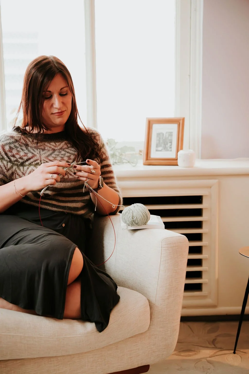 A woman with long dark hair knitting with yarn and knitting needles on a cozy beige sofa in a well-lit room.