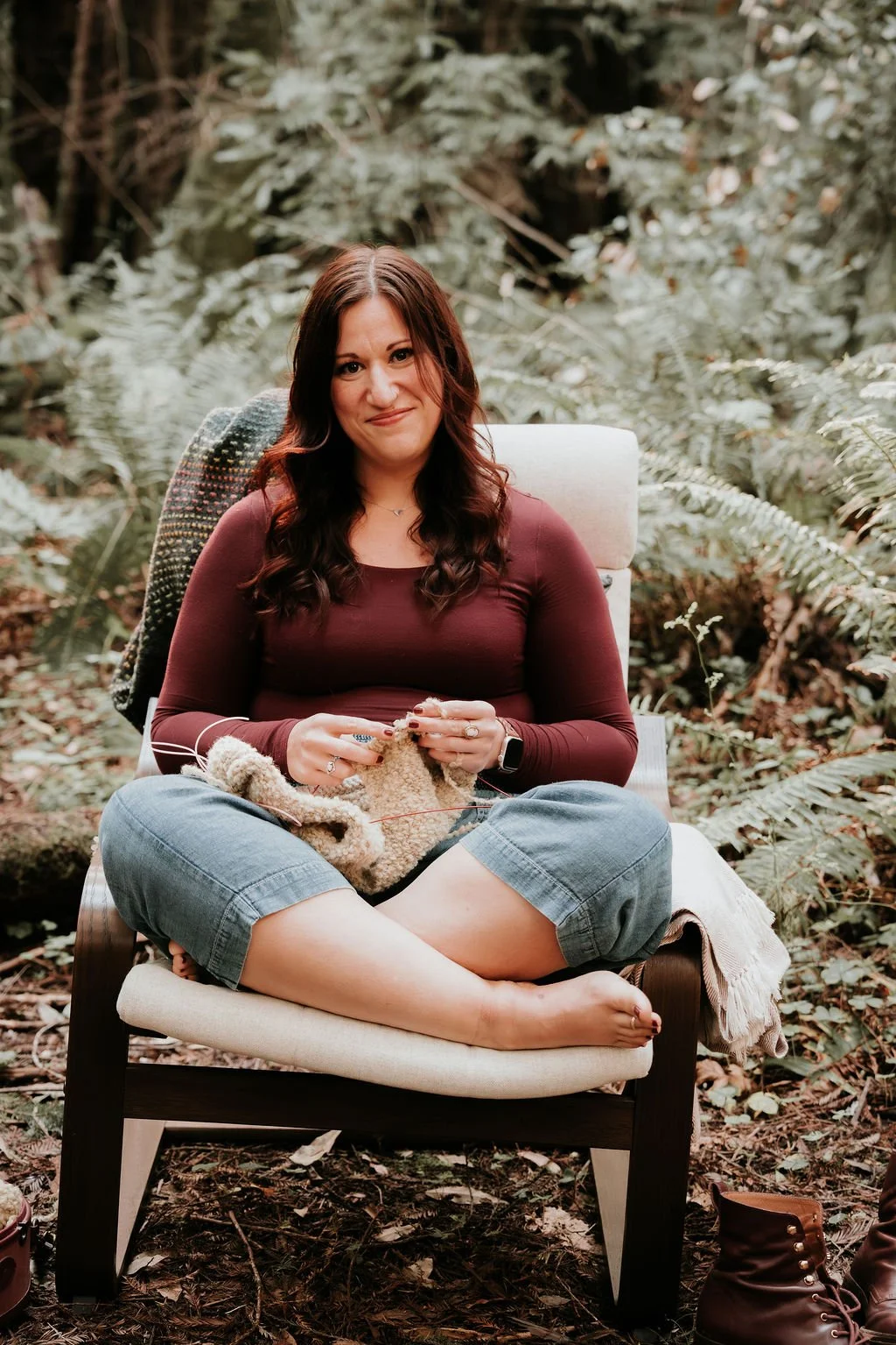 A woman with long brown hair sitting cross-legged in a forest, knitting with beige yarn, smiling at the camera.