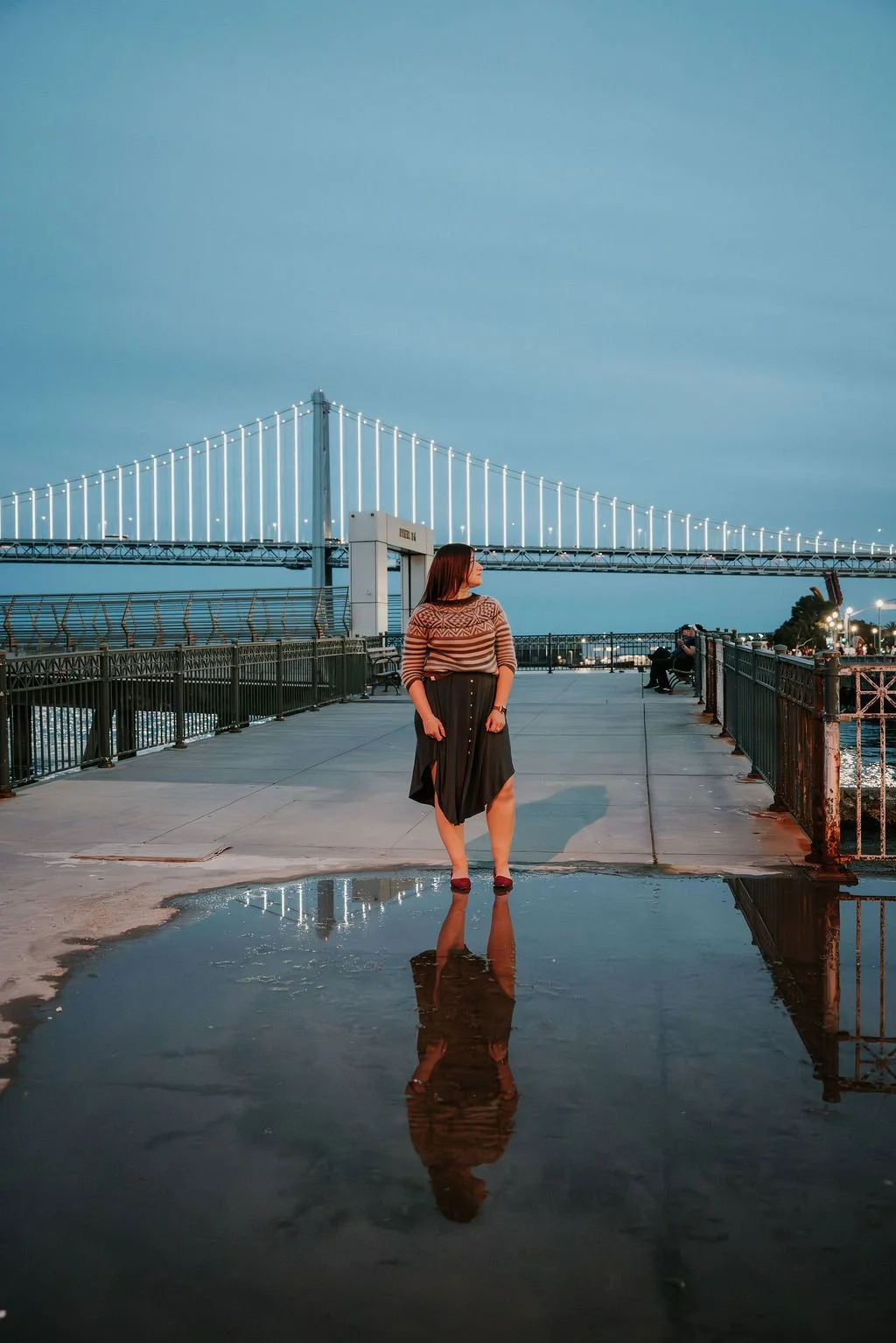 A woman standing on a waterfront promenade with a large suspension bridge illuminated in the background during dusk. Her reflection is visible in a puddle on the ground.