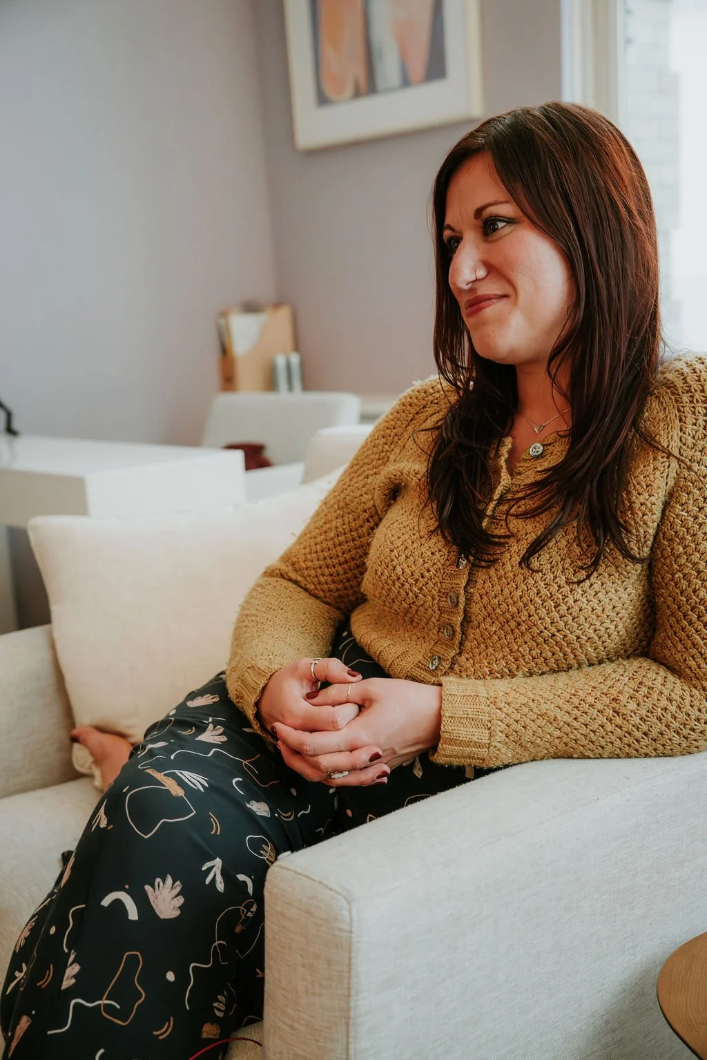A woman with long, brown hair, wearing a mustard-colored knit sweater, sitting on a beige couch with a pillow. She has a gentle smile and is clasping her hands. The background shows a gray wall with artwork and a white side table.