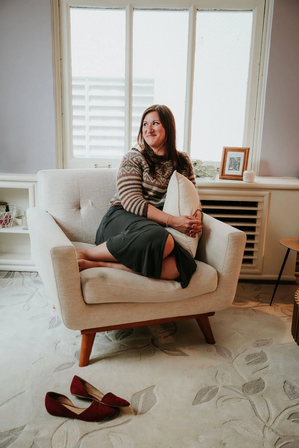 A woman sitting on a beige armchair holding a pillow in a room with a large window, picture frame, and decorative items.