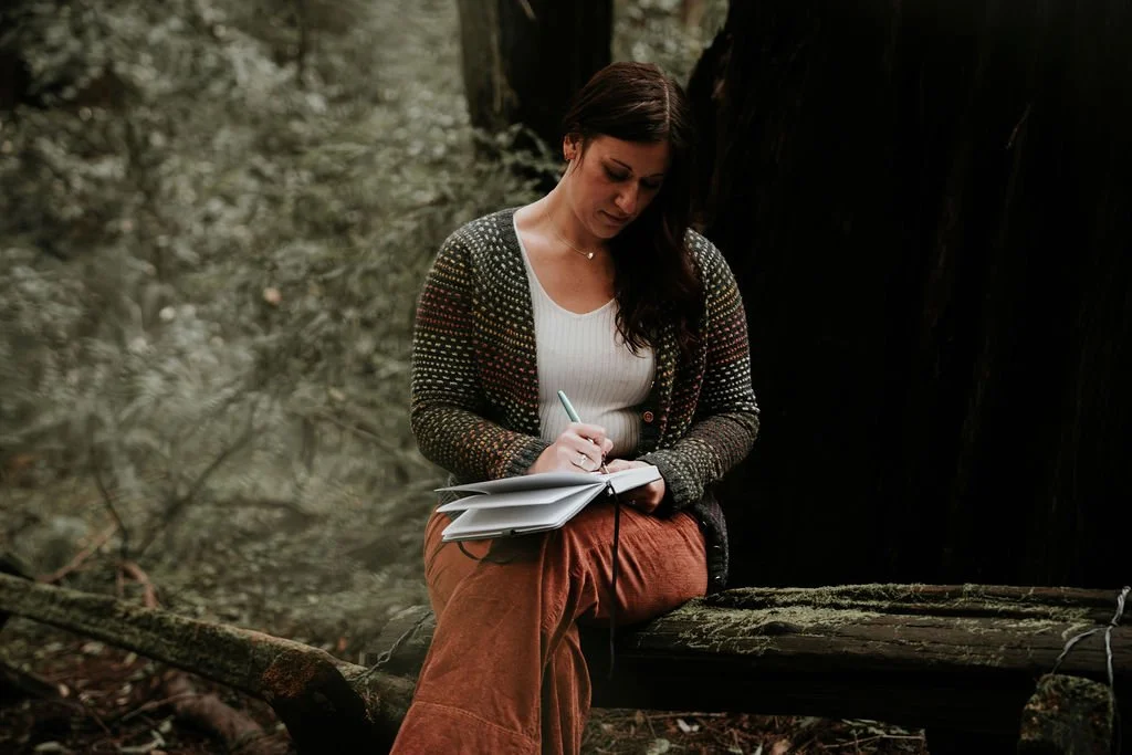 Woman sitting on a tree trunk in a forest, writing in a notebook.