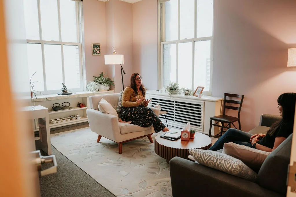 Two women sitting in a well-lit living room having a conversation, with large windows, a white sofa, a dark armchair, and various decorations.