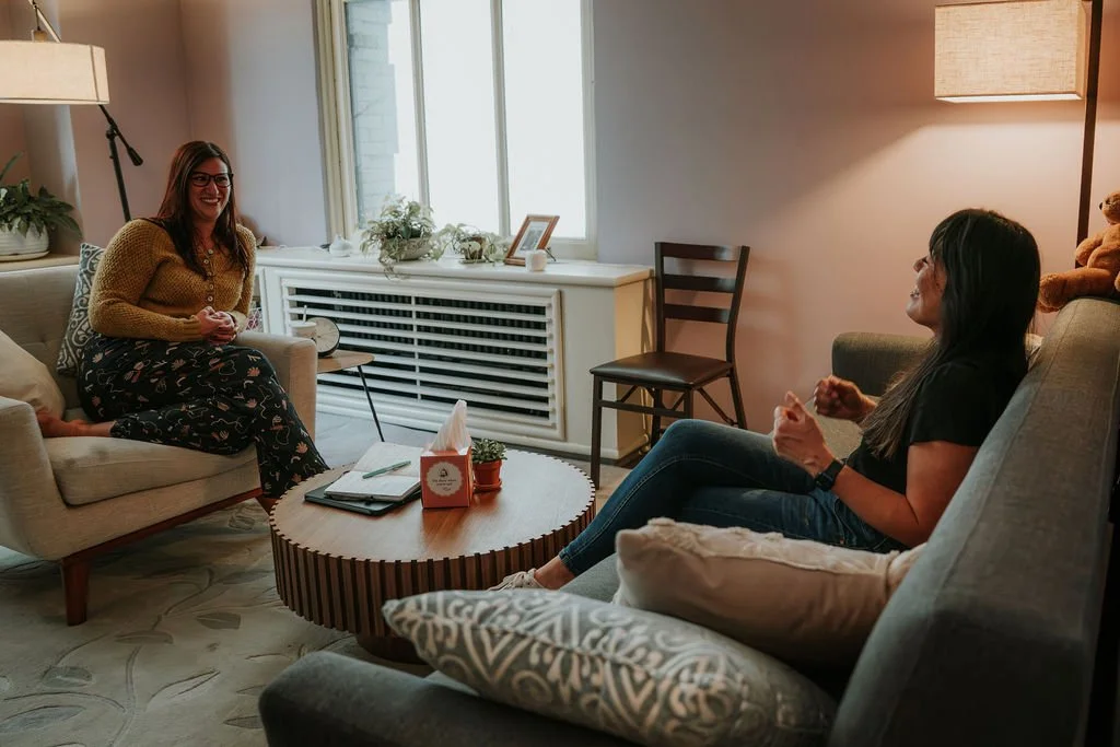 Two women are sitting in a living room having a conversation. One woman is on a light-colored sofa with glasses and a yellow sweater, while the other is on a darker sofa wearing a black shirt. There is a round coffee table with notebooks and a box of tissues in front of them. The living room has a window, decorative plants, and lamps providing warm lighting.