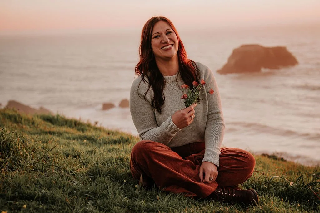 A woman sitting on the grass near the ocean, holding a small bouquet of flowers with a rock formation in the background at sunset, smiling at the camera.