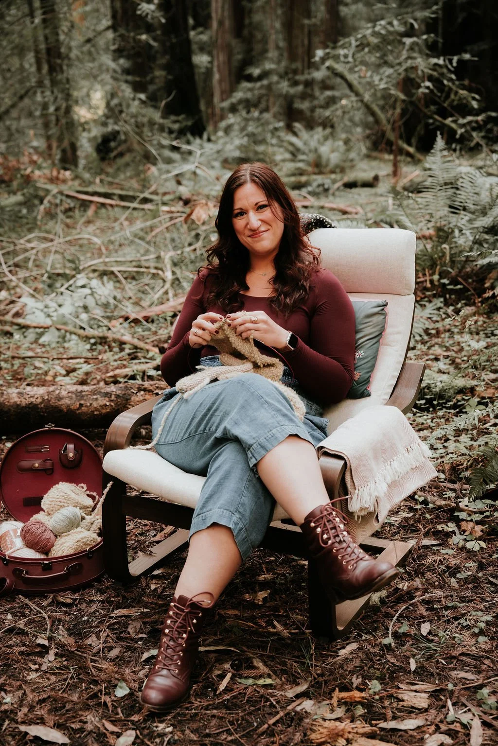 Woman sitting on a wooden chair with a pillow and knitted blanket, knitting in a forest surrounded by fallen leaves and plants, with a red knitting yarn case and yarn balls on the ground.