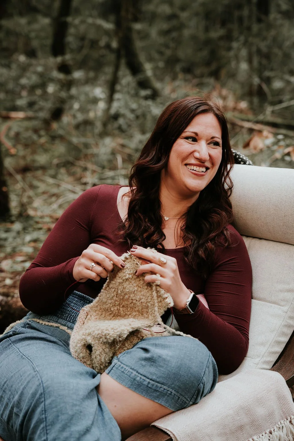 A woman with long dark wavy hair, smiling, sitting outdoors on a cushioned chair in a wooded area, knitting with beige yarn, wearing a maroon long-sleeve shirt and blue denim shorts.