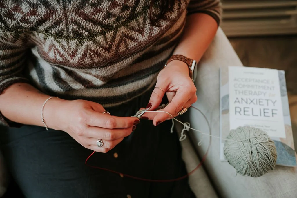 A woman knitting with yarn, with a book titled 'Acceptance, Commitment Therapy for Anxiety Relief' and a ball of yarn on a table nearby.
