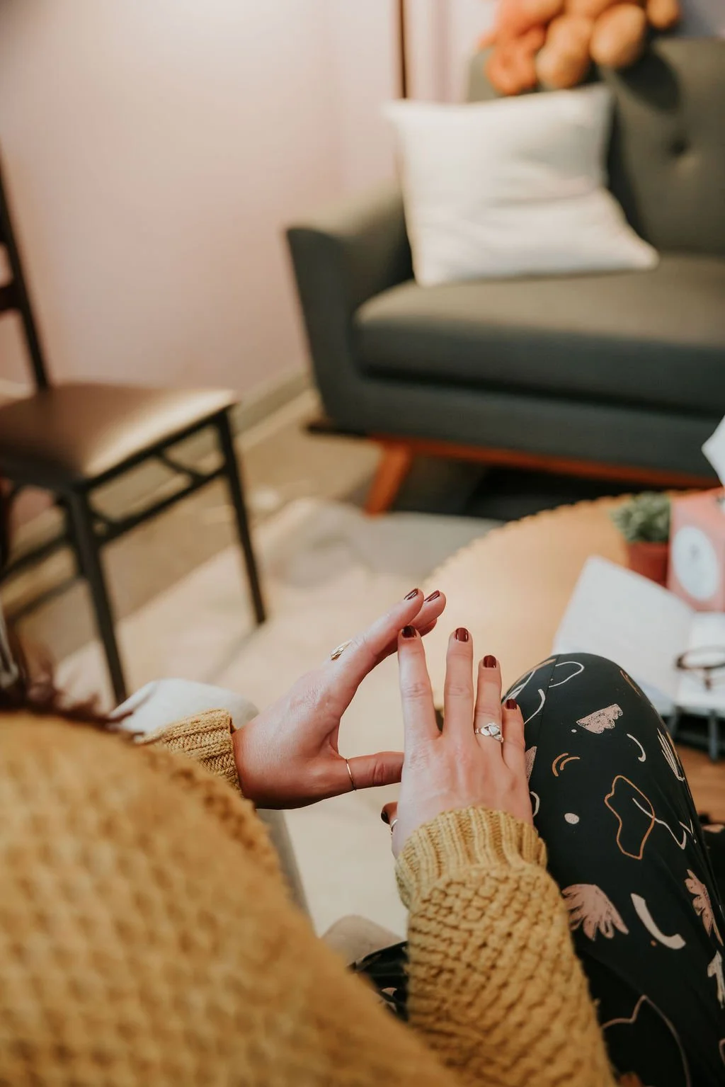 Close-up of a woman wearing a mustard knit sweater, sitting on a beige chair. She has her hands raised, with manicured nails painted dark and rings on her fingers. In the background, there is a gray sofa with a white pillow, a wooden chair, a small pink plant pot, and a table with items on it.