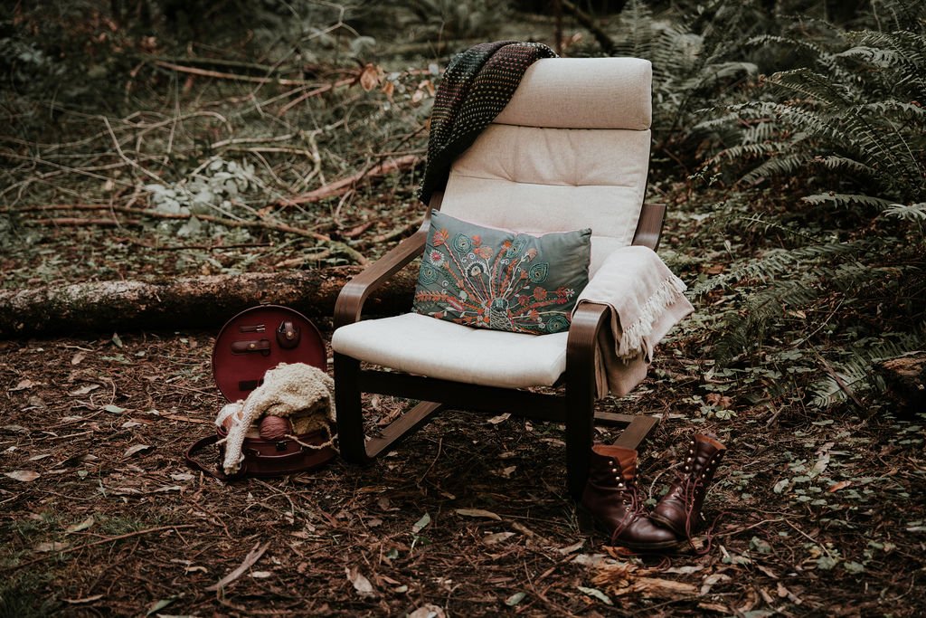 A white outdoor rocking chair with a beige cushion, a decorative pillow, a black blanket, and a cream-colored throw blanket is placed in a forest. Next to the chair, there are brown boots, a round red bag with a bottle inside, cream yarn, and pink yarns, all on the forest ground covered with leaves and twigs.