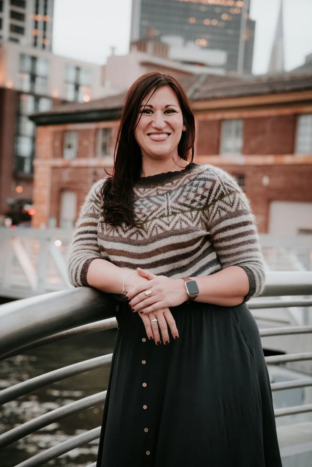 A woman with dark hair smiling, wearing a patterned sweater and a black skirt, standing by a metal railing near water with city buildings in the background during dusk.