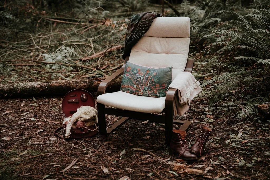 An outdoor scene with a white cushioned armchair surrounded by ferns and fallen branches. A colorful pillow, a cream blanket, a pair of brown boots, and a small round case with earbuds and a phone are placed around the chair.