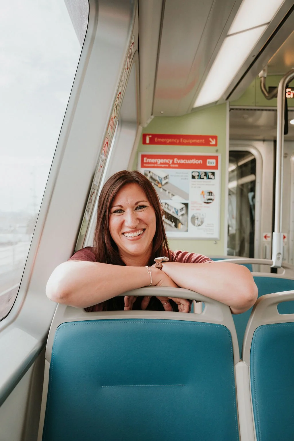 A woman with brown hair smiling and leaning on the seat back on a train, with emergency signage and a window in the background.