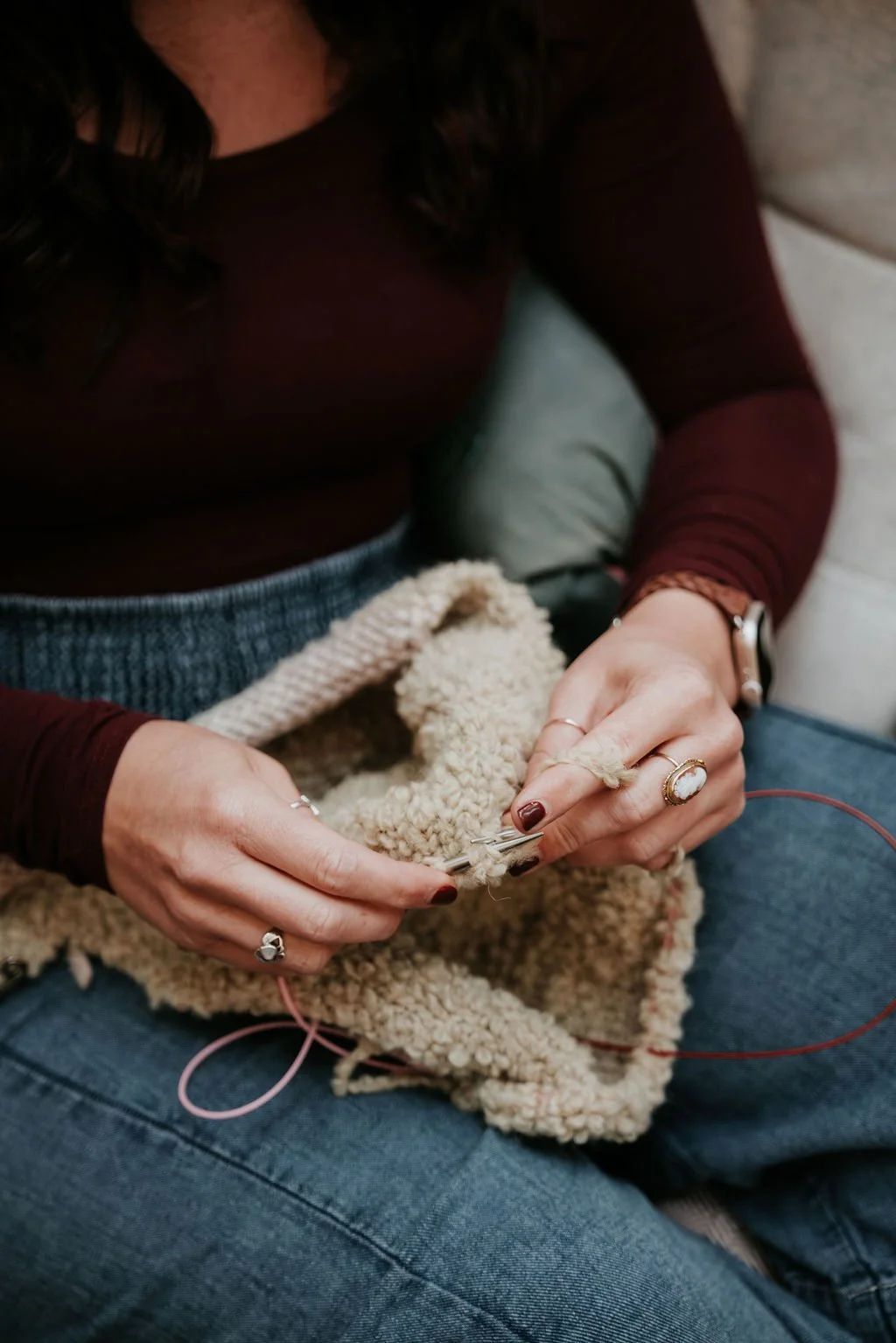 A woman knitting with cream-colored yarn on her lap, wearing a maroon long sleeve shirt, blue jeans, multiple rings, a watch, and a bracelet.