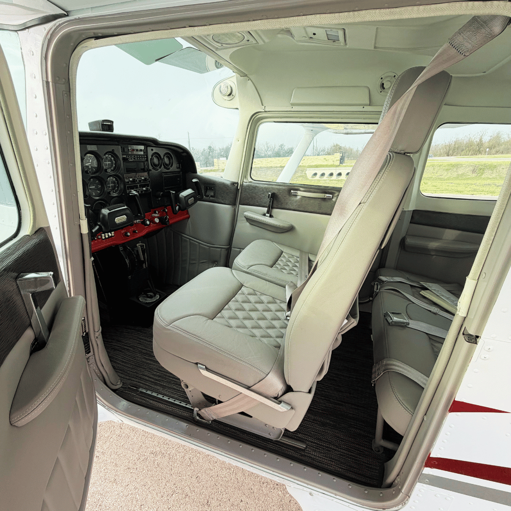 Interior view of a small airplane cockpit with two gray quilted seats, a control panel, and a window showing a grassy field outside.