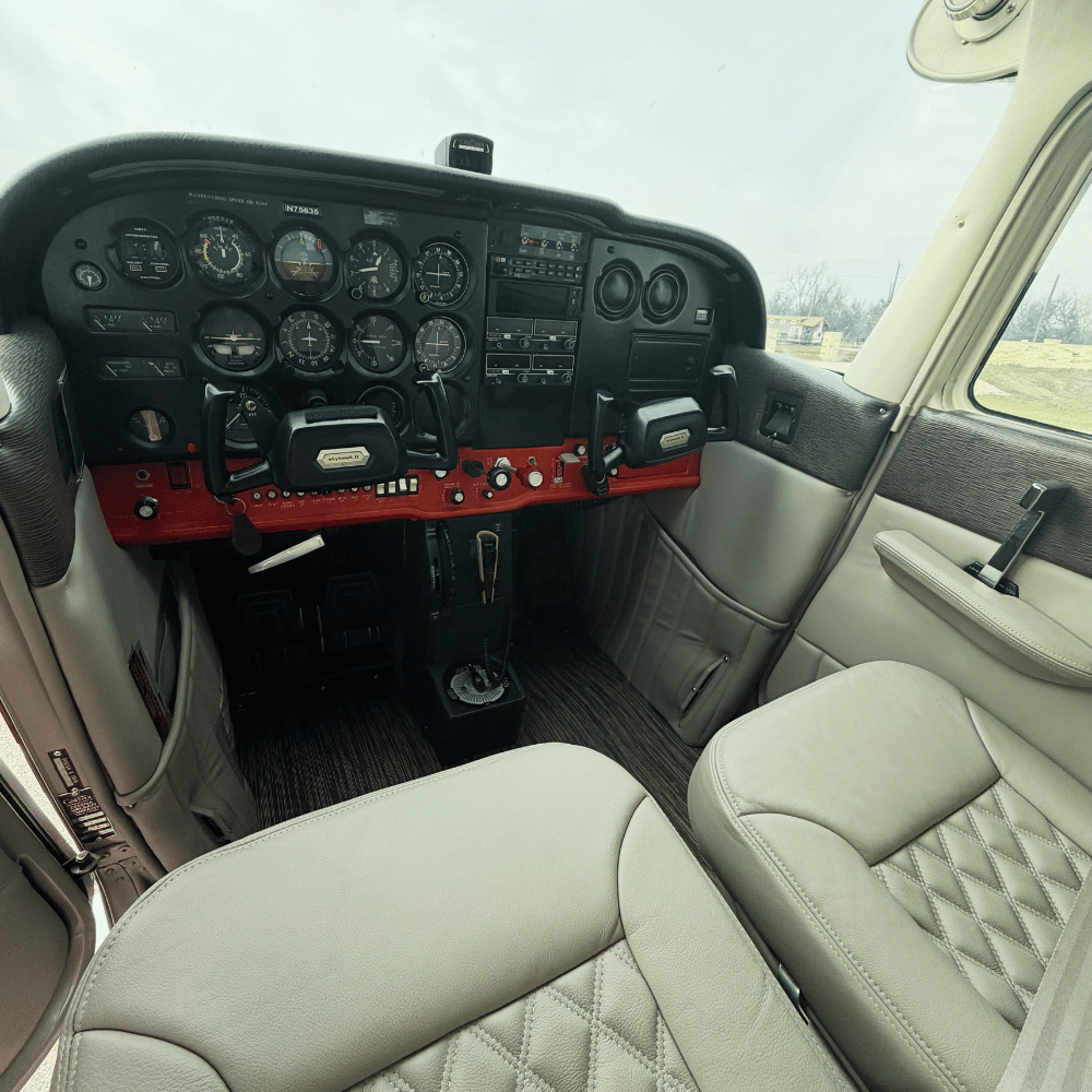 Interior of a small airplane cockpit showing various gauges, controls, and a dual yoke steering system, with cream-colored cushioned seats and a window.